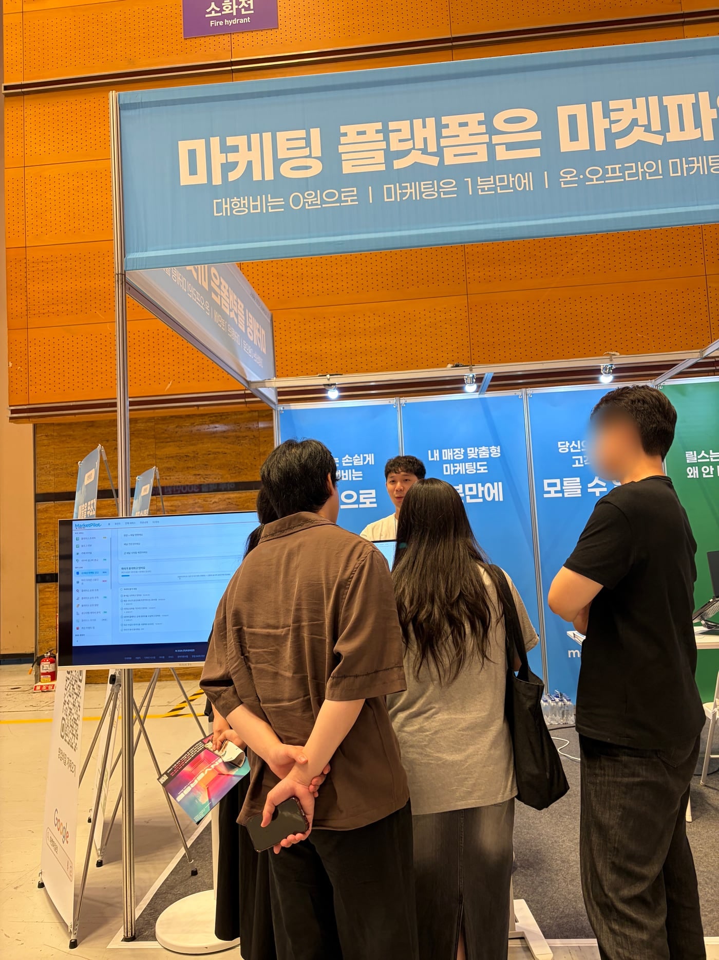코엑스 창업박람회의 마켓파일럿 조립부스. 위에 상호간판, 칸막이 벽면 그래픽, 시연 모니터, 상담하는 관람객들(관람객 얼굴은 흐리게 처리)